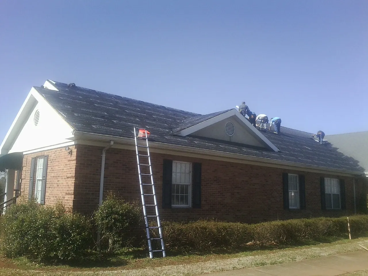 Crew tearing shingles off a brick ranch roof with a ladder set — Metal Roof Repair work by our crew in Knotts Island