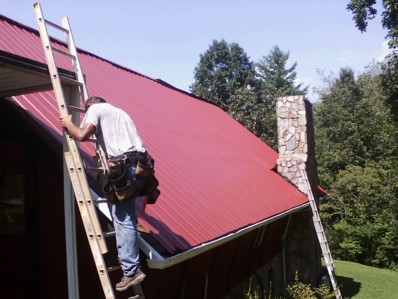 Roofer on a ladder beside a red metal roof and stone chimney — Soffit & Fascia Repair by Roofing Brothers in Knotts Island